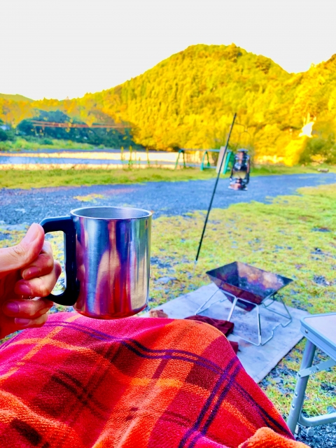 紅葉の山を前に、ひざ掛けの上でマグカップを持つキャンプの朝