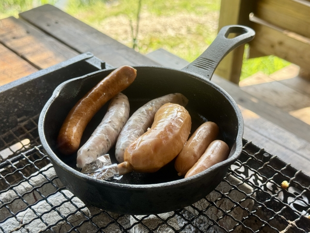 焚き火台の網にのせたスキレットで焼いているソーセージ
